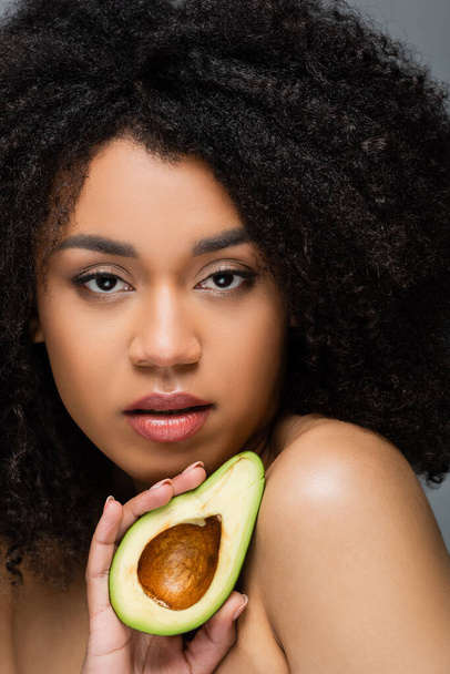pretty african american woman with natural makeup and perfect skin holding cut avocado isolated on grey - Foto, afbeelding