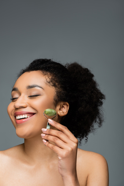 happy african american woman with perfect skin and natural makeup holding jade roller isolated on grey - Fotó, kép