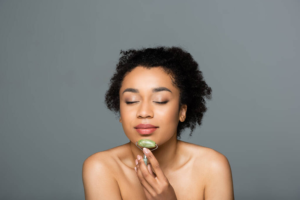 young african american woman with naked shoulders holding jade roller isolated on grey - Photo, Image