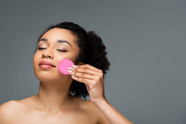 young african american woman with bare shoulders and closed eyes applying face foundation with sponge isolated on grey - Фото, зображення