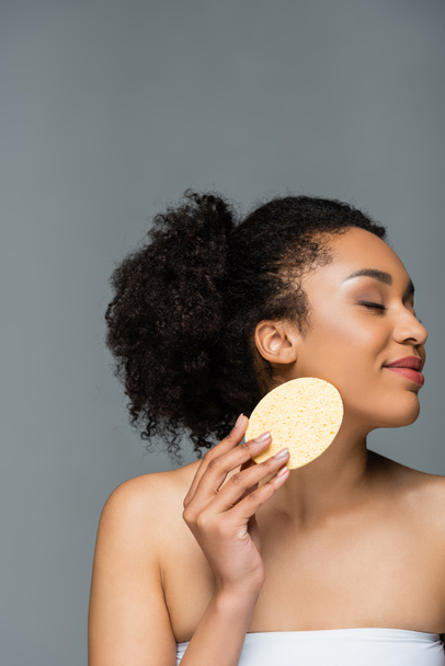 pleased african american woman with closed eyes wiping face with cosmetic sponge isolated on grey - Fotografie, Obrázek