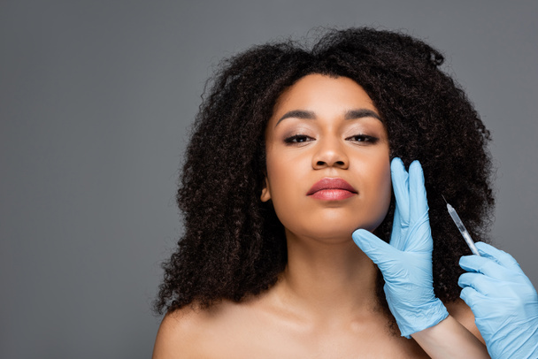 κοσμητολόγος σε γάντια λατέξ κρατώντας ένεση ομορφιάς κοντά στο πρόσωπο της αφρικανικής αμερικανικής γυναίκας που απομονώνονται σε γκρι - Φωτογραφία, εικόνα