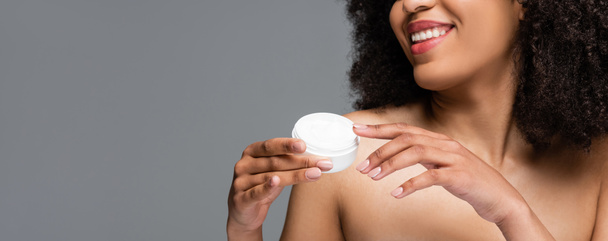 partial view of smiling african american woman with naked shoulders holding cosmetic cream isolated on grey, banner - Fotografie, Obrázek