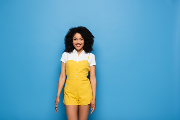 trendy Afrikaans amerikaanse vrouw in geel jumpsuit glimlachen op camera op blauw - Foto, afbeelding