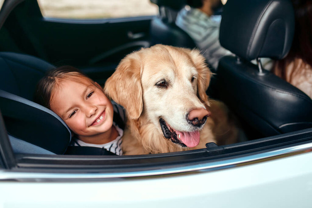 Cute little girl child in a car seat protected by seat belts together with her friend dog labrador going on weekend. Leisure, travel, tourism. - Photo, image
