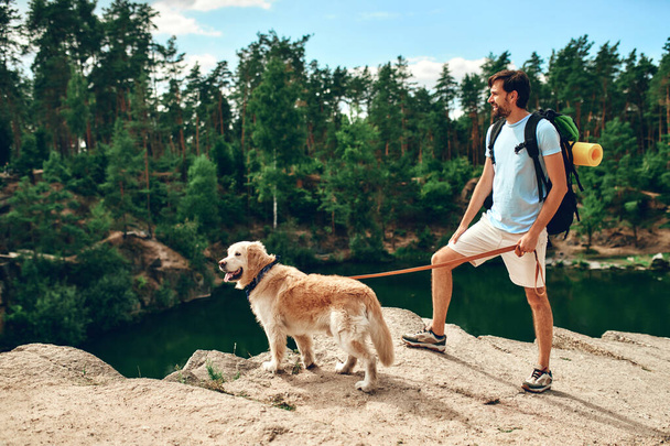 Чоловік з рюкзаком і собакою-лабрадором стоїть на скелястому піку, дивлячись на річку і ліс. Кемпінг, подорожі, піші прогулянки
. - Фото, зображення