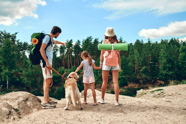 A family with backpacks and a labrador dog stands on a rocky peak, looking at the river and the forest. Camping, travel, hiking. - 写真・画像