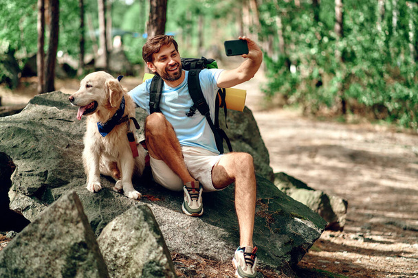 A male hiker with a backpack and a labrador dog makes a selfie on the phone while sitting on a stone in the forest. Camping, travel, hiking. - Foto, imagen