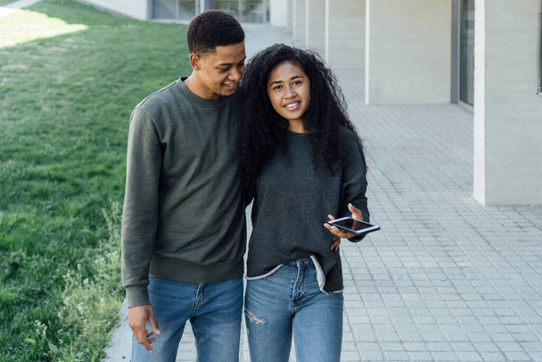Jovem casal negro caminhando abraçado ao usar um telefone celular ao ar livre. - Foto, Imagem