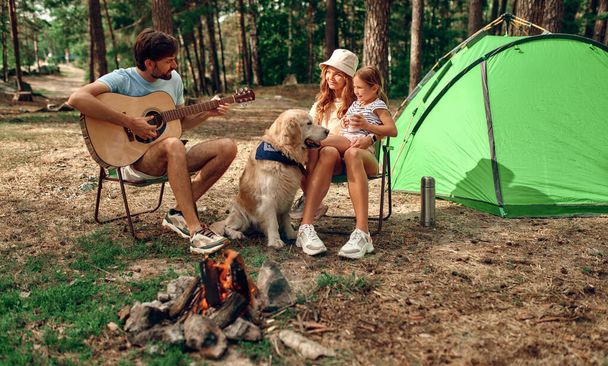 テントの近くのキャンプファイヤーに子供とラブラドール犬の若い家族が座って、松林の中でギターを演奏します。キャンプ、レクリエーション、ハイキング. - 写真・画像