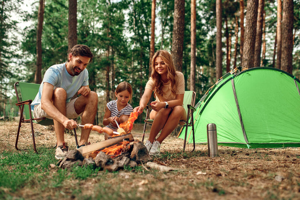 Szczęśliwa rodzina z dzieckiem na pikniku siedzieć przy kominku w pobliżu namiotu i grillować grilla w sosnowym lesie. Kemping, rekreacja, turystyka. - Zdjęcie, obraz