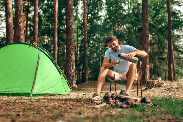Un hombre se sienta cerca de una tienda de campaña junto a una fogata y vierte café o té de un termo en un bosque de pinos durante el fin de semana. Camping, recreación, senderismo. - Foto, Imagen