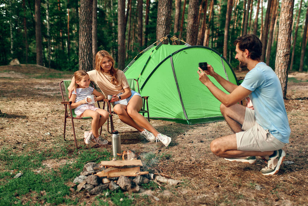 Szczęśliwa rodzina siedząca przy ognisku w pobliżu namiotu w sosnowym lesie na weekend. Mężczyzna robi zdjęcie żonie i córce przez telefon. Kemping, rekreacja, turystyka. - Zdjęcie, obraz