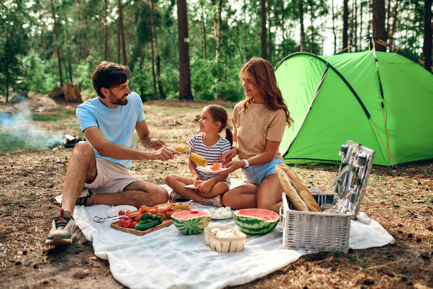 Happy family with a child on a picnic sitting on a blanket near the tent and eating food fried at the stake during the weekend in the forest. Camping, recreation, hiking. - Photo, Image