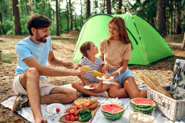 Happy family with a child on a picnic sitting on a blanket near the tent and eating food fried at the stake during the weekend in the forest. Camping, recreation, hiking. - Zdjęcie, obraz