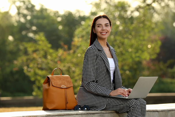 Beautiful young woman with stylish backpack and laptop on bench in park - Photo, Image