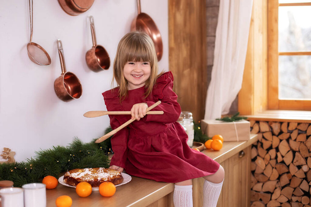 Lachend klein meisje zittend op het tafelblad met een houten keukenspatel in haar handen. Kind houdt vast en speelt met spatel kooktaart. gelukkig klein meisje in jurk koken kerstkoekjes thuis - Foto, afbeelding