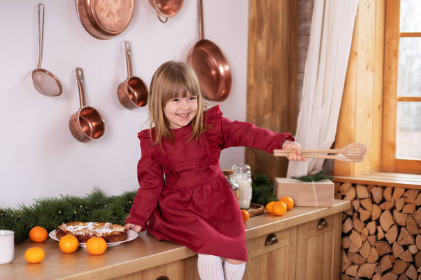 Dziecko trzyma i bawić się łopatką gotowania ciasto. Szczęśliwa dziewczynka w sukience gotowanie świąteczne herbatniki w domu. Uśmiechnięta dziewczynka siedząca na stole z drewnianą łopatką kuchenną w rękach - Zdjęcie, obraz