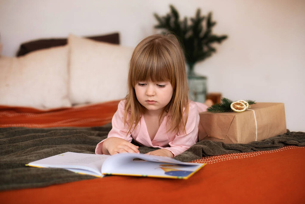 Kleines Mädchen im Bett liest ein Buch. Glückliches kleines lächelndes Mädchen im Schlafanzug mit Weihnachtsgeschenk liegt auf dem Bett. Kind hält Buch in der Hand und liest es, Kind betrachtet ihr Buch, liest interessantes Märchen. - Foto, Bild