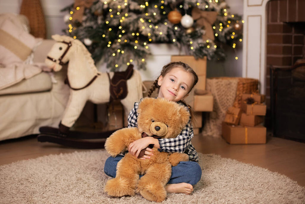 Happy little girl with pigtails sitting on living room floor and hugs big teddy bear toy. A child sits by Christmas tree with garlands and opens gifts at home. Concept new year and family holidays.  - Foto, imagen