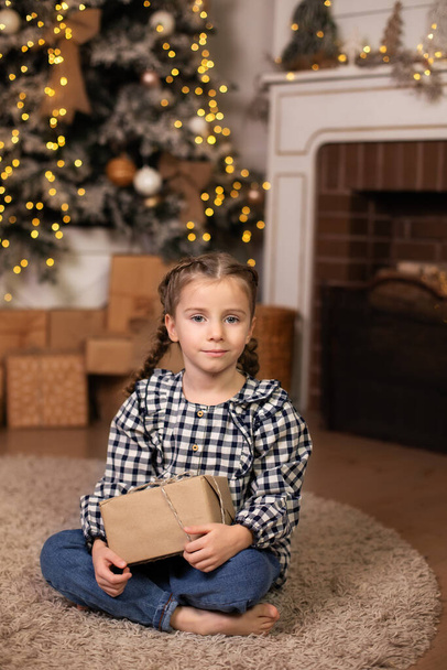  Happy little girl with christmas present at home. Small Girl holds gift near Christmas tree on lights background. Child opens Xmas gift in living room. New Year, winter holiday - Photo, Image