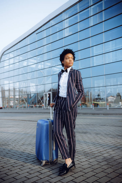 4k. Reisen, Digital. Charmante Afroamerikanerin im modern gestreiften Anzug steht an der Glaswand neben ihrer Reisetasche. Unternehmerin oder Studentin. Gleichgültigkeit. - Foto, Bild