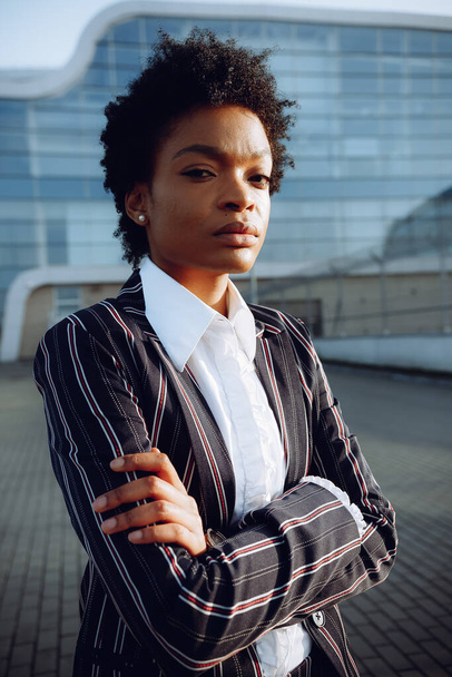 4kだ。エレガントなポーズでガラスの壁の現代的な縞模様のスーツの背景に魅力的なアフリカ系アメリカ人女性。実業家や学生。成功. - 写真・画像