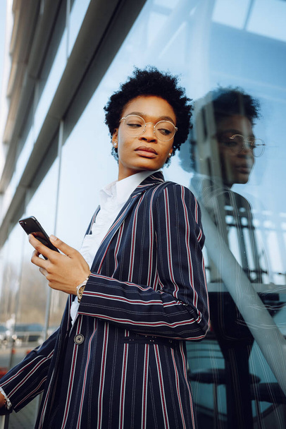 4k. Viaggio, digitale. Affascinante donna afroamericana in abito moderno a righe con bagagli e tenendo il telefono. Sta vicino al muro di vetro e aspetta il suo volo. Imprenditrice o studentessa. - Foto, immagini