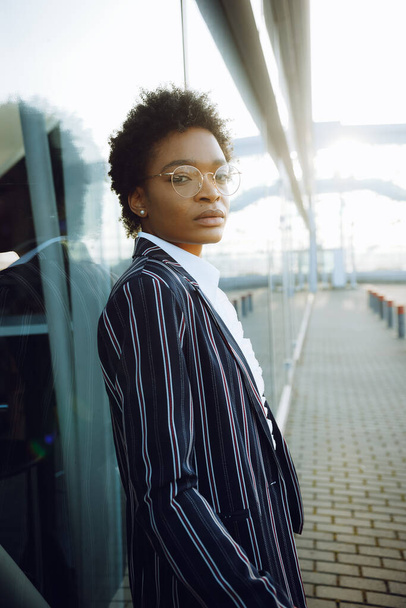 4k. Travel, Digital. Charming African American woman in elegant striped suit. Stands by the glass wall and looks in camera. Businesswoman or student. Thoughts. Sky. - Photo, image