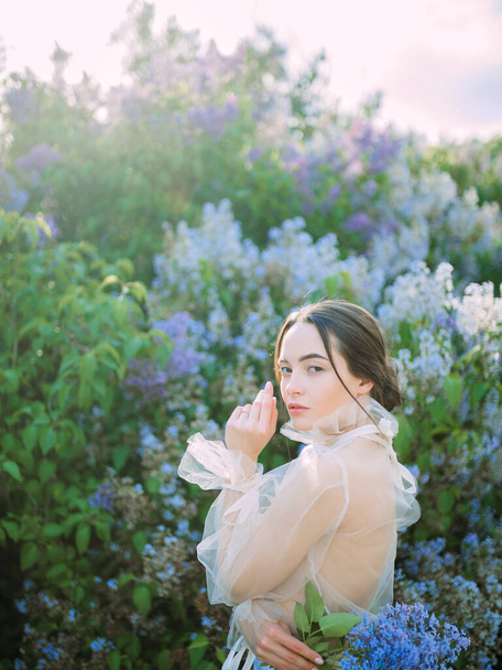 Young woman walks in garden with branch of lilac in her hand against background of blooming shrubs. - Photo, Image