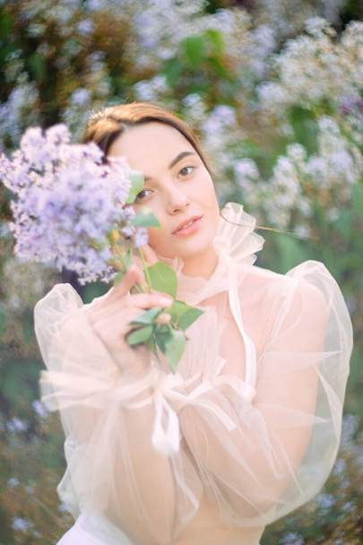Portrait of young woman in garden with branch of lilac in her hand against background of blooming shrubs. - Photo, Image