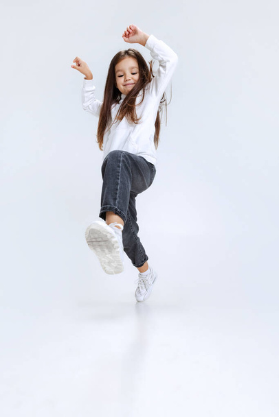 Full-length portrait of little cute girl, kid wearing hoodie with jeans steps, jumps isolated over white studio background. - Fotoğraf, Görsel