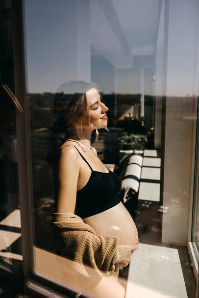Zwangere vrouw thuis bij het raam in het zonlicht, glimlachend. - Foto, afbeelding