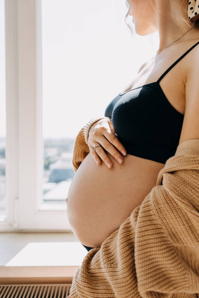 Primer plano de una mujer embarazada parada junto a la ventana con luz suave. - Foto, imagen