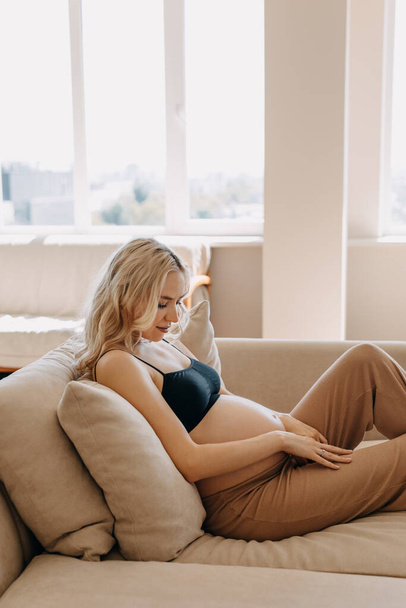 Mulher grávida vestindo roupas acolhedoras em casa, deitada em um sofá em um interior leve em casa. - Foto, Imagem