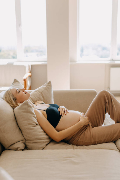 Беременная женщина в уютной домашней одежде, лежит на диване в светлом интерьере, держит руки на животе. - Фото, изображение