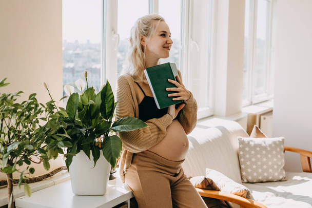 Pregnant woman wearing cozy home wear, holding a book, smiling. - Valokuva, kuva