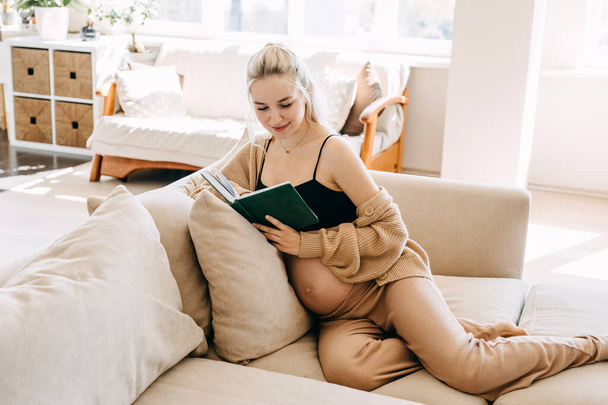 Pregnant woman wearing cozy home wear, sitting on a sofa at home, reading a book. - Foto, Imagem