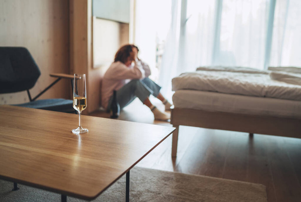 Бокал вина на столе с расфокусированной грустной женщиной, сидящей на полу в спальне с бутылкой алкоголя. Проблемы психического здоровья и алкоголизма. - Фото, изображение
