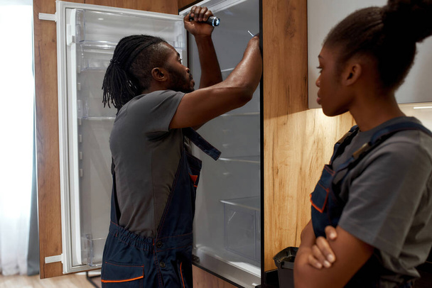 Gender inequality. Female repair service worker standing by male technician repairing fridge - Foto, Imagen