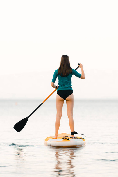 Young girl on a sup board with a paddle in her hand - shot from the back, calm surface of the sea, copy space - Foto, imagen