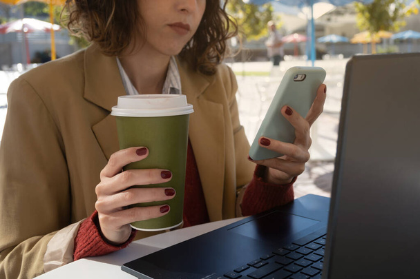 Nahaufnahme bis zur Unkenntlichkeit latina Frau, die am Computer arbeitet, während sie am Telefon schaut und Kaffee trinkt, mit Kopierplatz - Foto, Bild