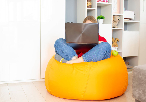Eine junge Frau sitzt auf einem orangefarbenen Sitzsack-Stuhl im Zimmer und arbeitet zu Hause mit einem Laptop. Fernarbeitskonzept, freiberuflich. - Foto, Bild