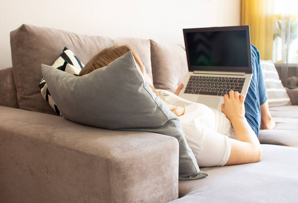 若い女性が部屋のソファに横になり、自宅のノートパソコンを使って仕事をしています。リモートワークの概念、フリーランス. - 写真・画像