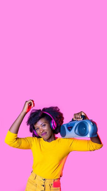 Young black woman listening music dancing holding vintage boombox and wearing wireless headphones on 16:9 vertical copyspace banner - Photo, Image