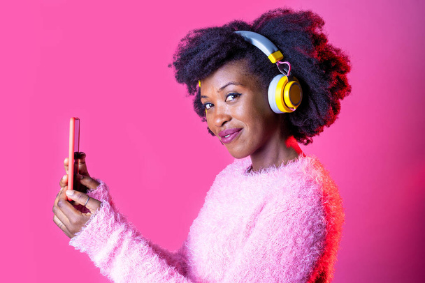 Молодая черная женщина слушает музыку беспроводные наушники принимая селфи держа смартфон изолированный розовый фон - Фото, изображение