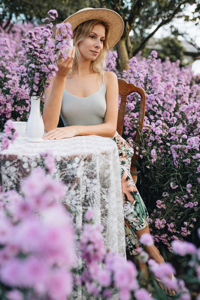 Young blonde woman sitting at the table with cup of tea in flower garden in Mon Jam, Thailand - Photo, Image