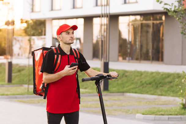 Male courier food delivery with red thermal backpack walks street with electric scooter uses smartphone navigate. Deliveryman worker employee deliver online order client customer at sunset. - Foto, Bild