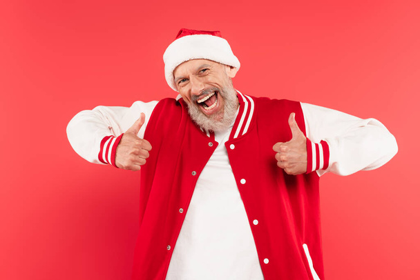 excitado hombre de mediana edad en sombrero de santa mostrando pulgares hacia arriba aislado en rojo - Foto, Imagen