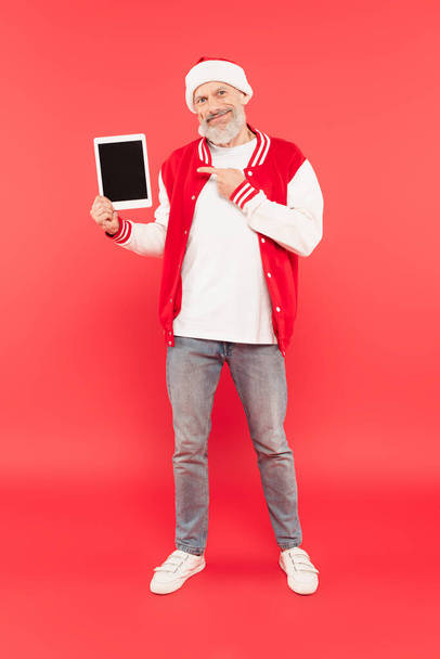 volledige lengte van gelukkig middelbare leeftijd man in santa hoed wijzend op digitale tablet op rood - Foto, afbeelding
